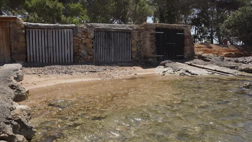Clear, calm water gently laps the shore of a secluded cove with rustic stone and wood boathouses