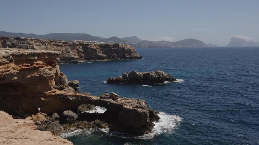 Scenic wide shot of a rugged Mediterranean coastline featuring a natural rock arch and blue waters on a sunny day