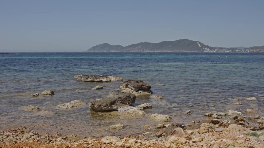 A picturesque view from a rocky shoreline overlooking the calm, clear turquoise sea with a mountainous island in the distance