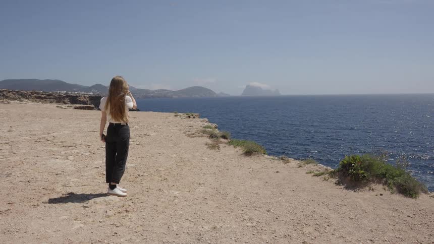 A young woman with long hair stands on a cliff overlooking the ocean and takes a photo on her phone