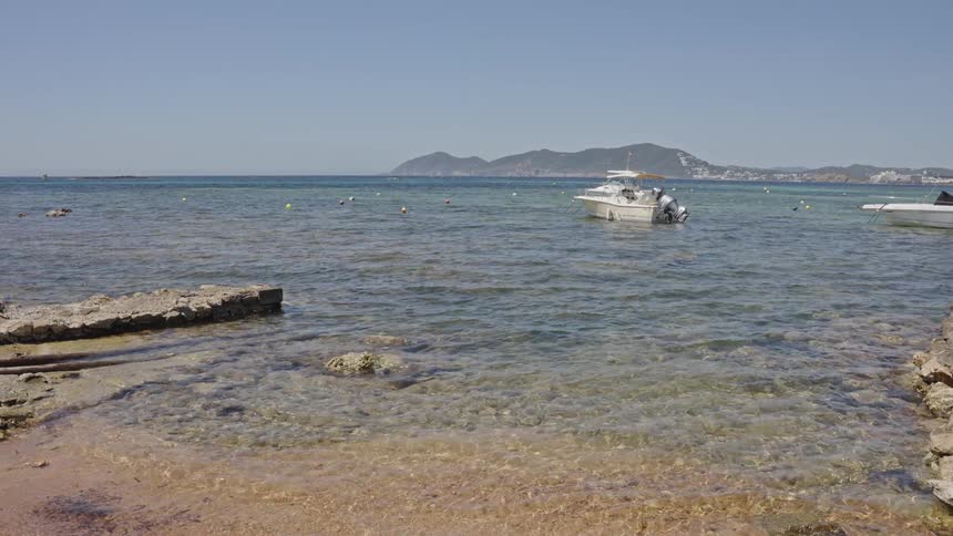 Boats float on tranquil turquoise waters near a sandy and rocky shore with distant hills under a clear blue sky