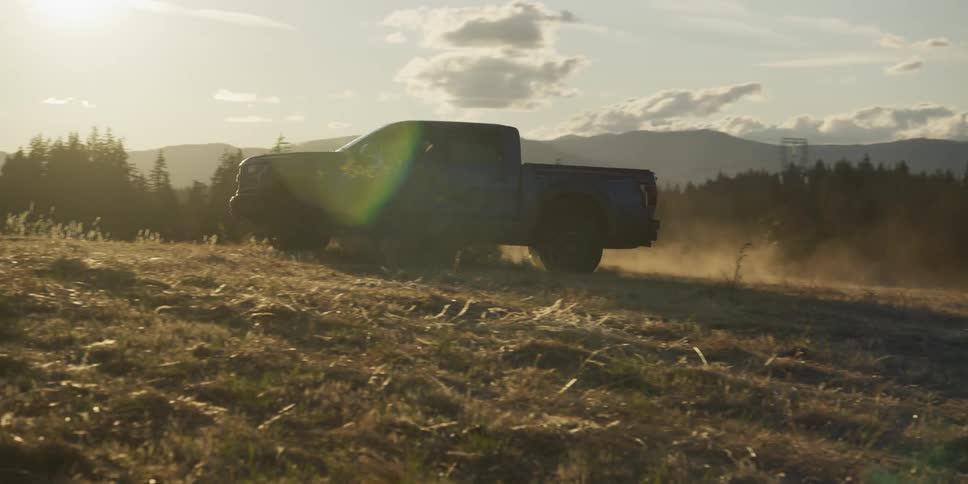 Majestic horses gallop in slow motion past a blue pickup truck in a field at sunset, before the truck drives off-road through the dusty mountain landscape