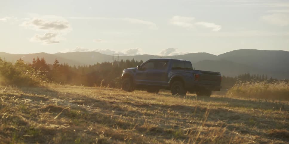 A modern pickup truck drives off-road through a golden mountain landscape as wild horses and a cowgirl gallop past into the sunset
