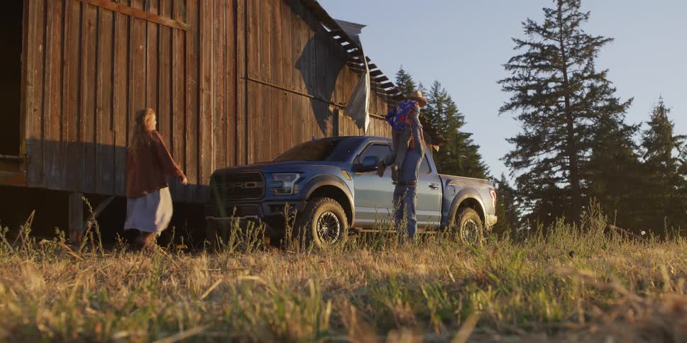 A family reunion unfolds as a boy dressed as a superhero cowboy jumps out of a blue truck to greet his father, with his mother following, all set against a rustic barn at sunset