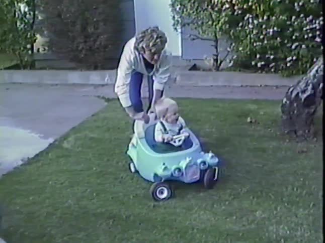 A mother pushes her happy baby in a blue toy car across the green lawn while another toddler watches