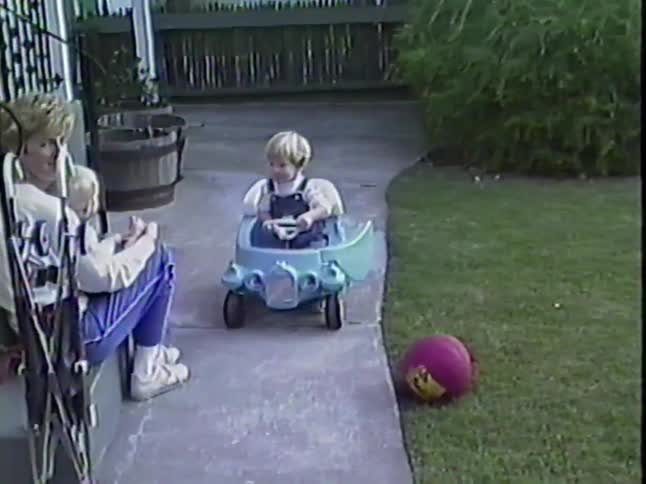 A mother sits on the front steps with her baby while her toddler son plays happily in a blue toy car on the sidewalk