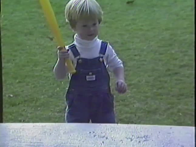 A young toddler in overalls plays with a toy baseball bat and ball in a grassy backyard in this vintage home video