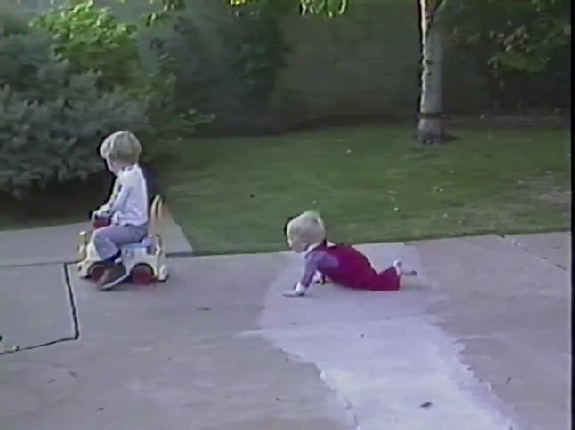 A baby in red overalls crawls across a backyard patio as their toddler sibling watches from a ride-on toy