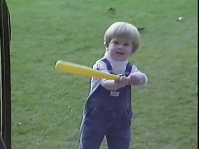 Vintage home video footage of a cute little boy in overalls swinging a toy baseball bat in a grassy yard