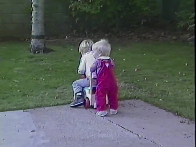 Two young siblings play with a ride-on toy car in their backyard in this authentic 1987 home video