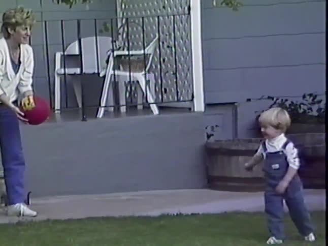 A young boy in overalls throws a red ball to his mom in the backyard, captured in nostalgic slow-motion home video footage