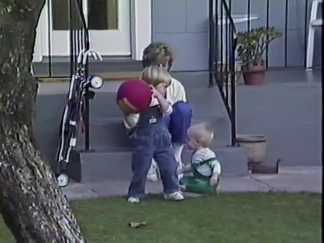A vintage home movie shows a mother watching her two young sons play with a ball in their front yard