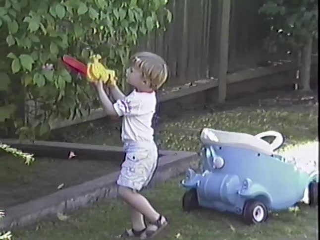 Retro VHS footage captures a cute toddler pretending to be a lumberjack with his toy chainsaw in the backyard