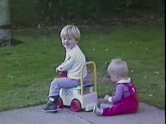 A toddler sits on a ride-on toy train as his baby brother falls down behind him on the sidewalk