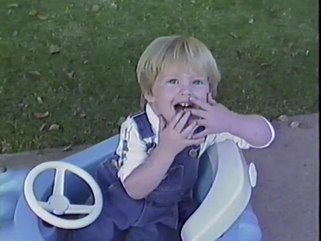 A joyful toddler in a blue toy car laughs at the camera before getting his face messy with a chocolatey snack