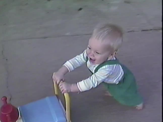 A joyful barefoot toddler in green overalls happily learns to walk by pushing a toy train on a sidewalk past a chalk drawing that reads 'I love you'
