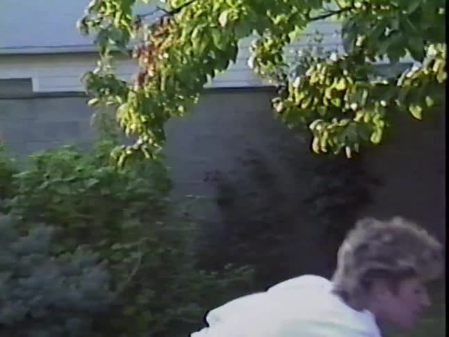 A toddler in green overalls plays with a push car on the patio while their mother in 80s attire watches and picks up a red ball