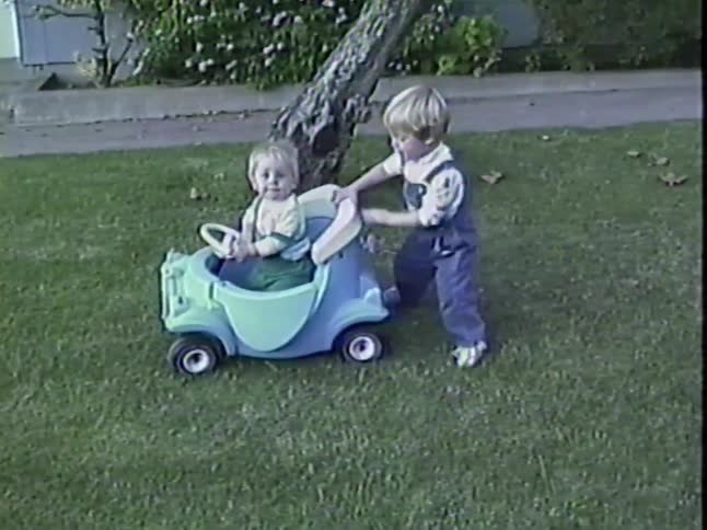 An adorable toddler pushes his baby brother in a toy car across the lawn in this nostalgic 1987 home video