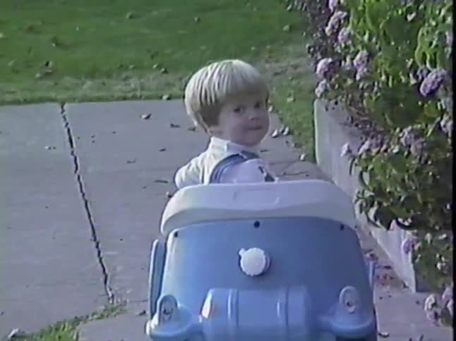 A cheerful toddler boy in overalls smiles while being pushed in a blue toy car on a sidewalk