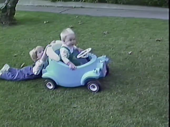 A vintage home movie shows a toddler pushing his baby brother in a toy car through the grass in their backyard