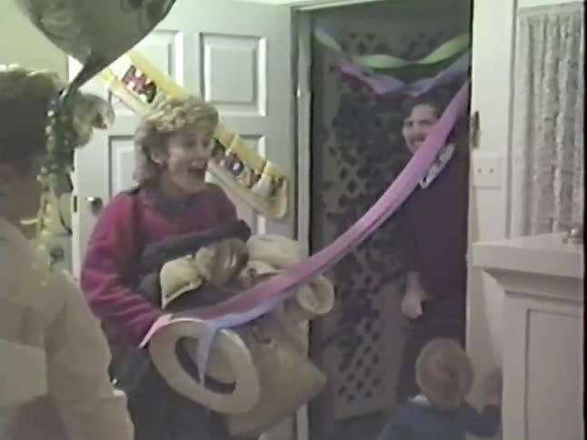 A woman carrying bags and a jacket is completely shocked as she walks into a surprise birthday party in this vintage home video
