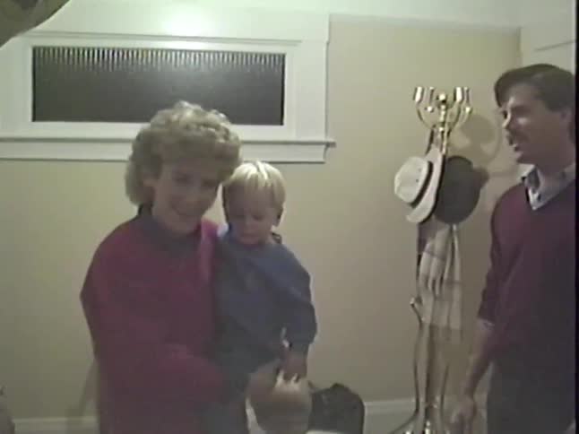 A smiling mother holds her young child while surrounded by family in a vintage home video from the 1980s