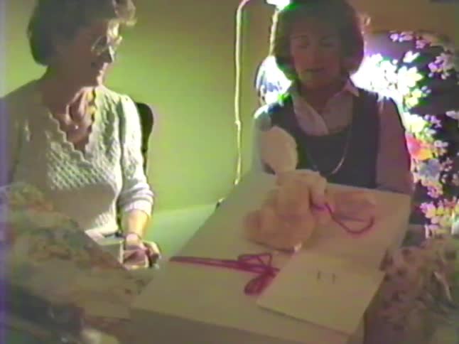 A vintage VHS home video captures two women talking at a baby shower with a large gift on the table