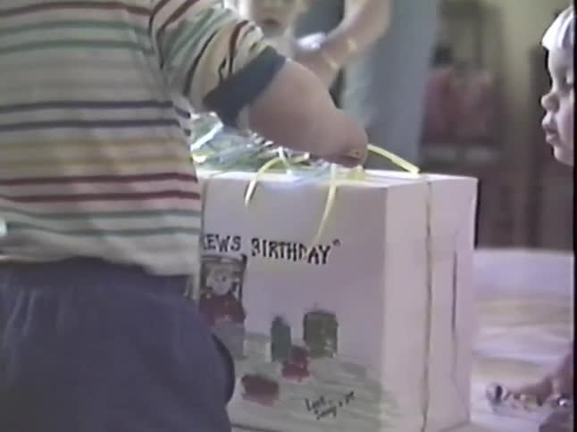 A nostalgic home video from the 1980s shows young children gathered around a large, hand-decorated birthday present