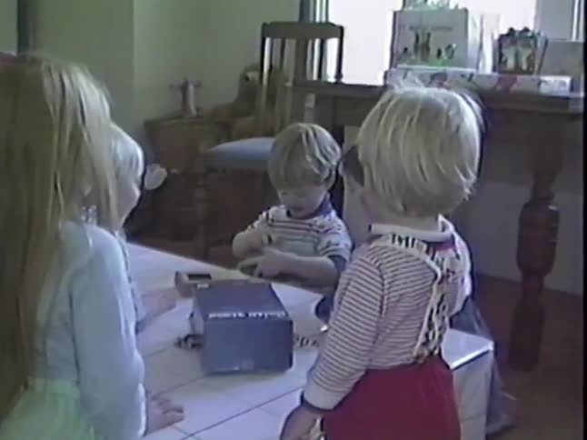 Vintage home video footage shows a toddler excitedly unwrapping a new toy car at his birthday party surrounded by his friends