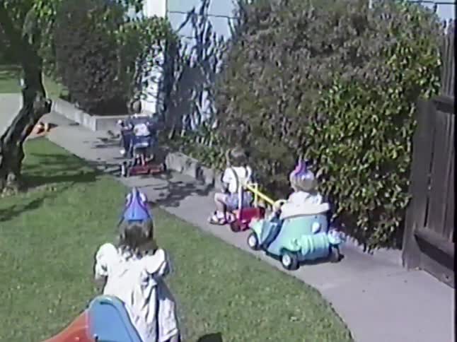 Vintage footage captures a parade of adorable toddlers in party hats riding their toy vehicles around a sunny backyard