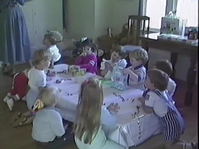 Adorable toddlers sit on the floor around a small party table, enjoying a celebration in this charming vintage home video