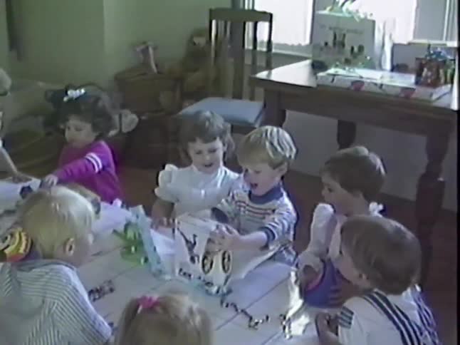Vintage home video footage shows a group of toddlers gathered around a table watching as a young boy opens his birthday presents