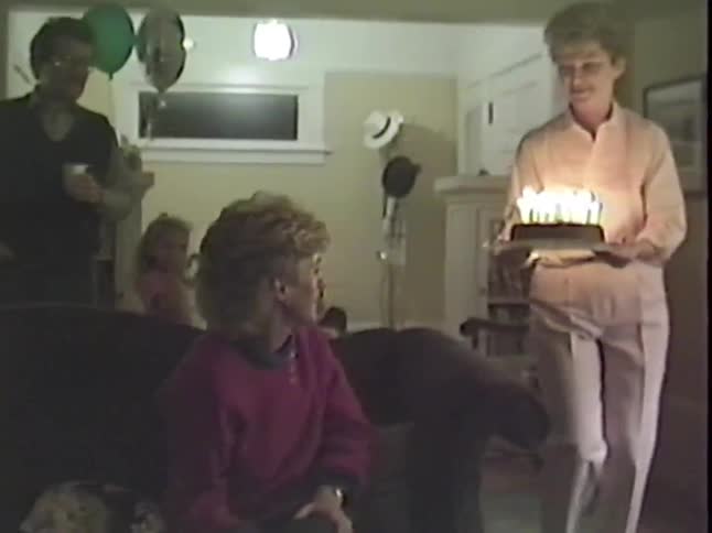 A woman brings a birthday cake with lit candles to another woman sitting on the couch while their family watches in this vintage home movie