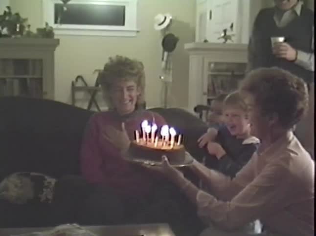 A woman laughs as she tries to blow out trick candles that keep relighting on her birthday cake in a nostalgic 1980s home video