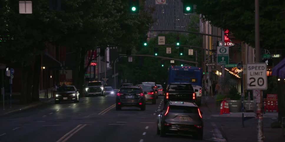 Slow zoom on a line of cars waiting at a red light on a city street at dusk, which turns green and traffic begins to move