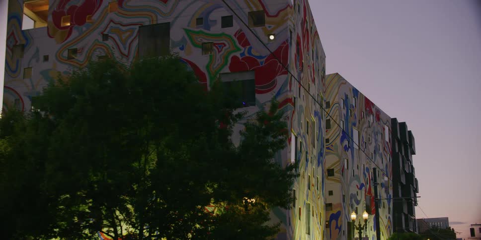 A slow tracking shot pulls back from a colorful abstract mural on a modern building at dusk, revealing the urban streetscape