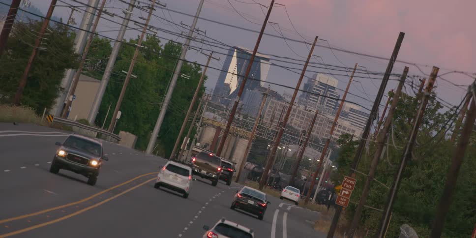 A slow zoom on a line of traffic commuting down an industrial road lined with telephone poles towards a city skyline at dusk