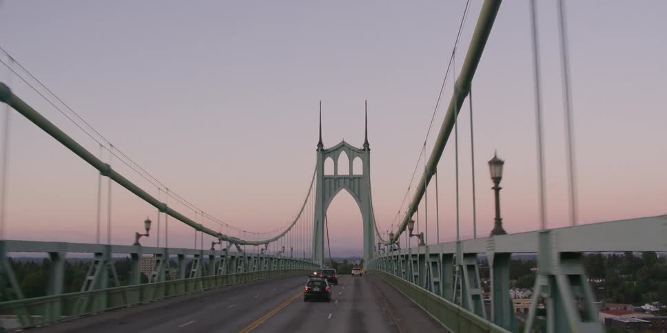 A serene aerial view shows cars commuting across the historic St. Johns Bridge against a pastel sunset sky in Portland, Oregon