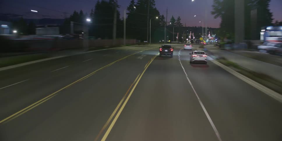 A point-of-view shot follows traffic on a multi-lane road at twilight, with oncoming cars creating mesmerizing light trails