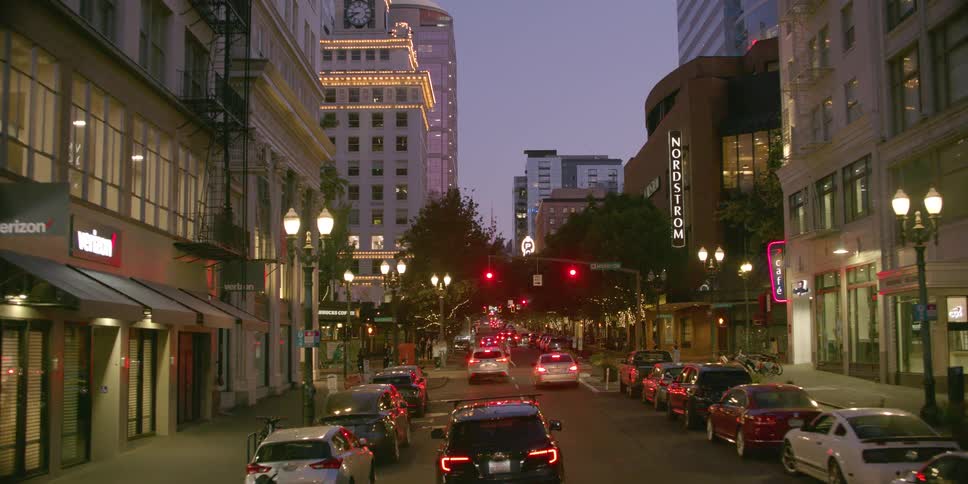 A slow, forward-moving view down a downtown city street at dusk, revealing illuminated buildings, traffic, and pedestrians