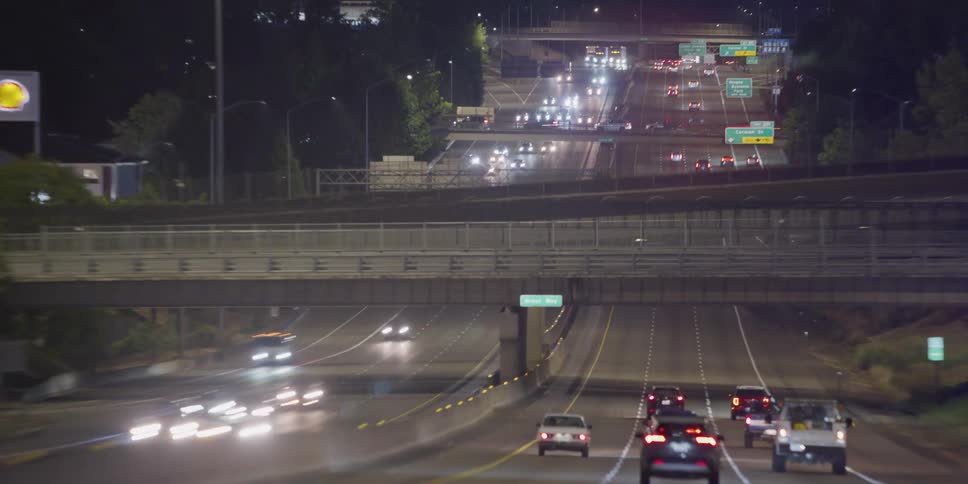 A slow zoom-in shot from a moving vehicle captures the flow of traffic on a busy multi-lane highway at night