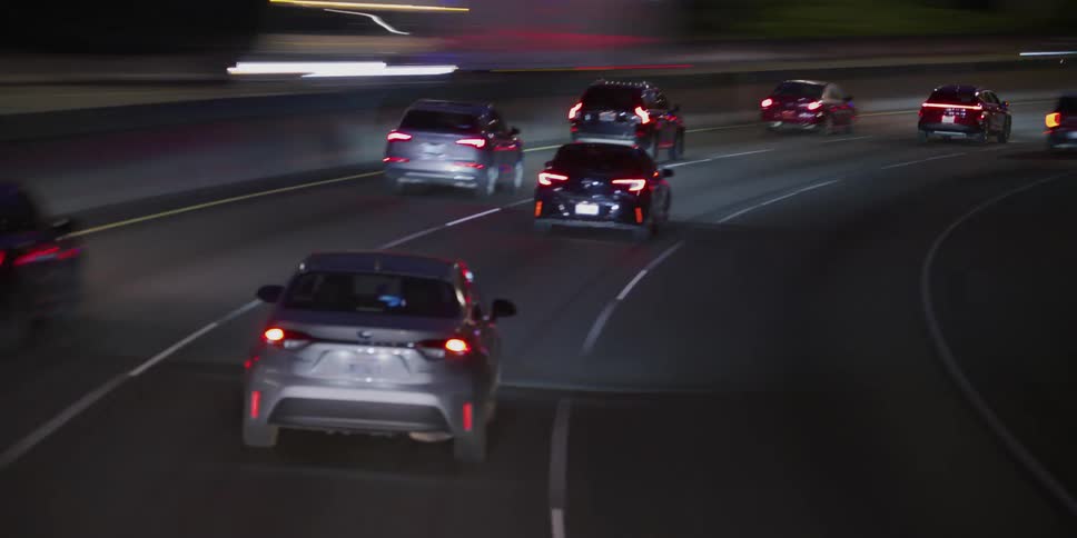 Following a steady stream of traffic down a curving multi-lane highway at night, with the headlights and taillights of cars illuminating the road