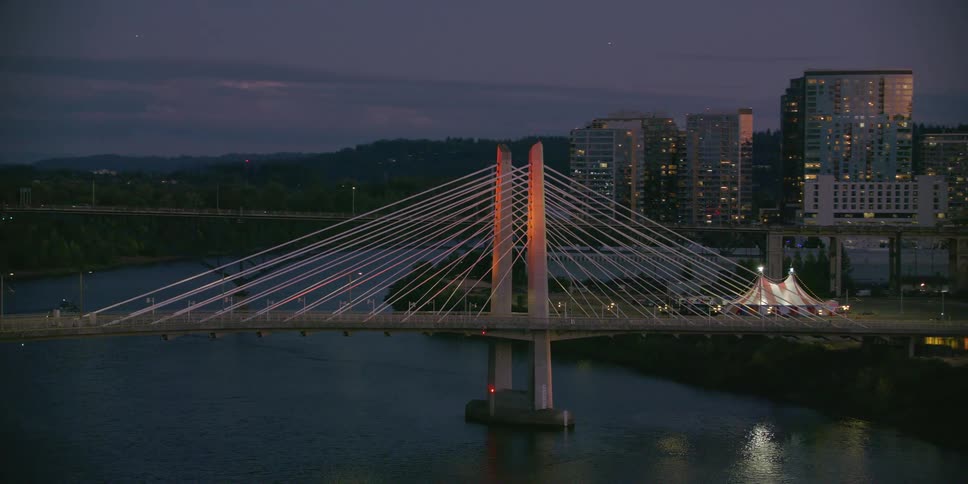 A slow aerial shot captures the beautifully lit Tilikum Crossing bridge spanning the Willamette River in Portland, Oregon at dusk