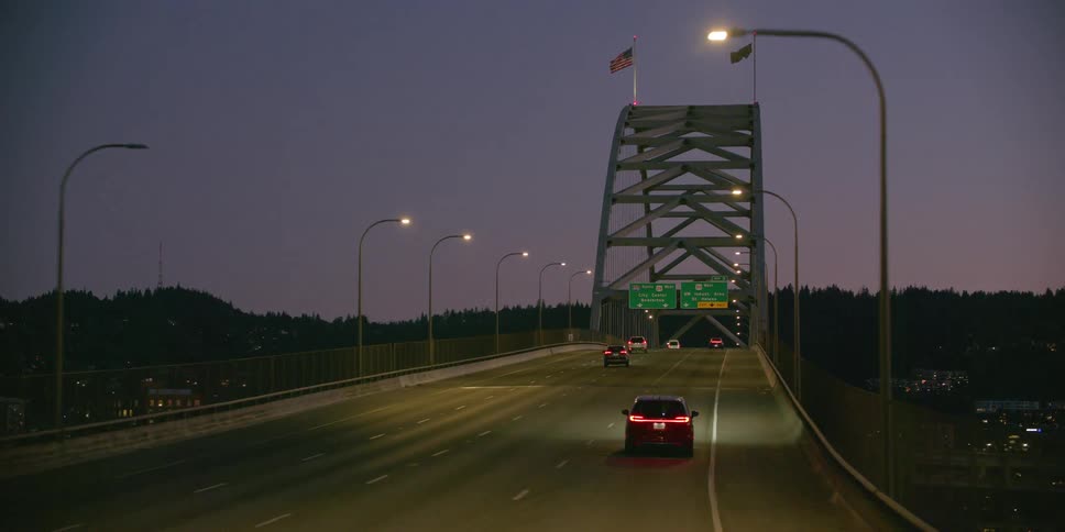 A smooth POV shot follows cars driving across a large, illuminated city bridge at dusk