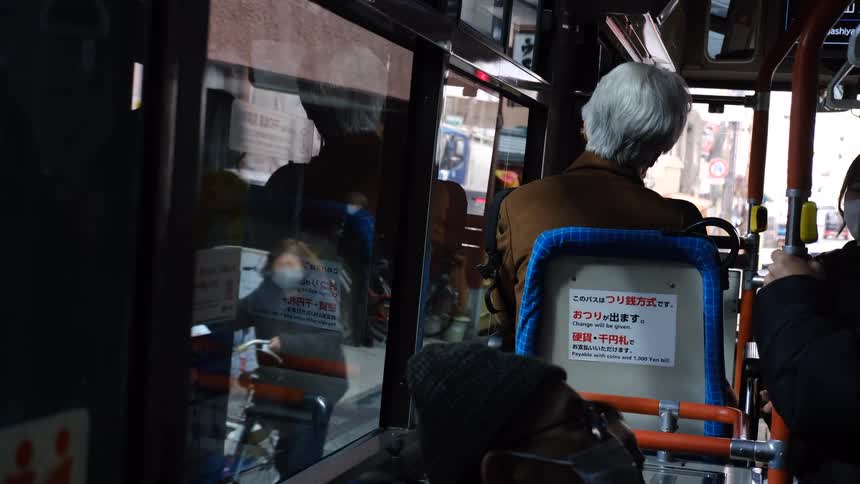 Slow motion view of a city street in Japan from inside a moving bus, with an elderly passenger in the foreground