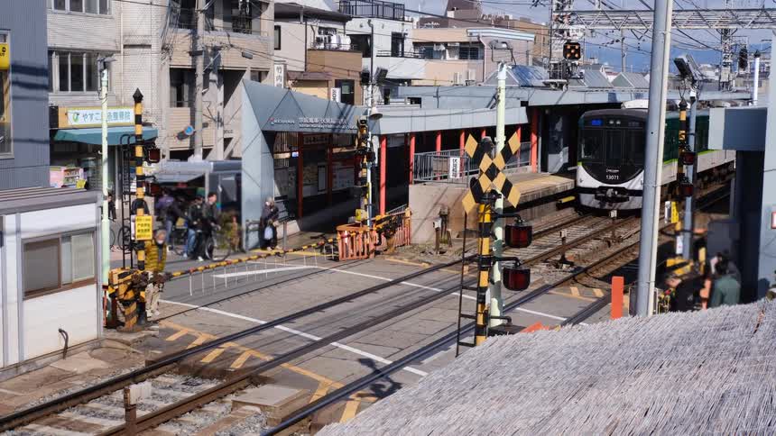 A Keihan electric train departs a station in urban Japan, and as it leaves, the railroad crossing gates lift for pedestrians to cross