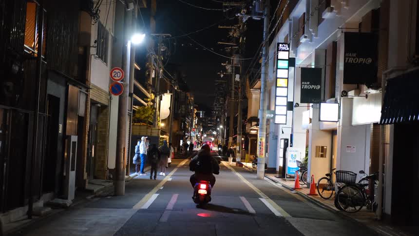 A person on a scooter drives down a quiet, narrow, illuminated street in a Japanese city at night