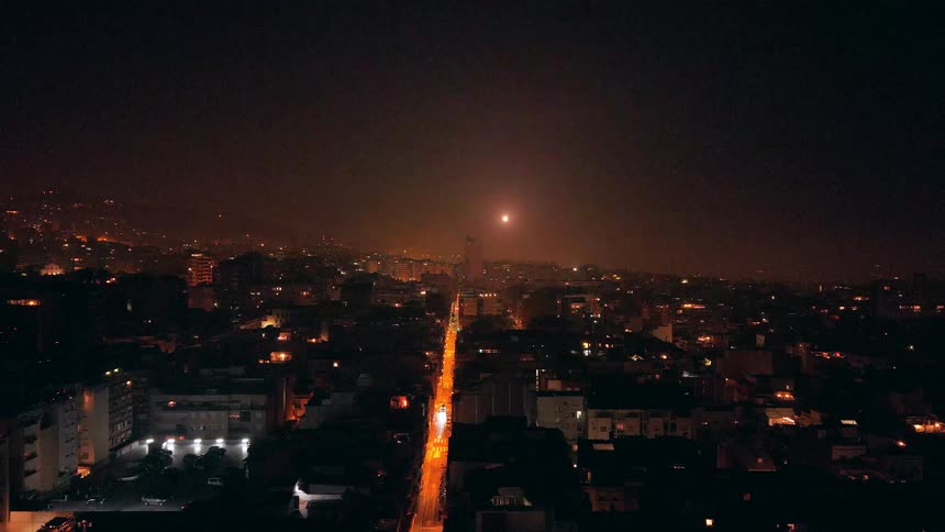 An expansive city glows with orange street lights as fireworks light up the dark night sky in a slow aerial reveal