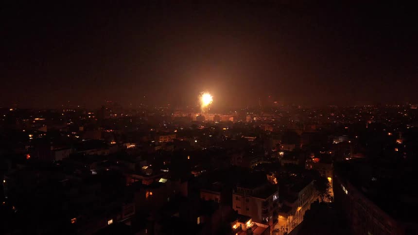 A static aerial shot captures a lone, colorful firework exploding over a vast, illuminated cityscape at night