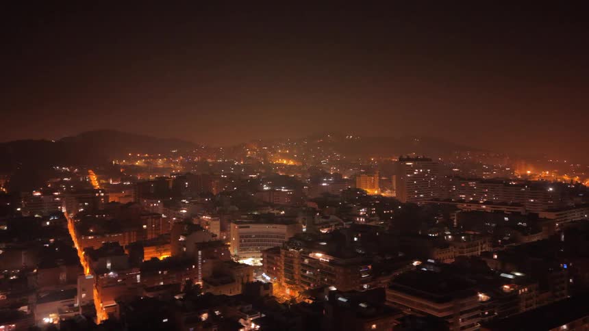 An aerial drone flies over a sprawling city illuminated by an orange glow on a hazy night